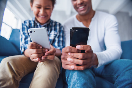 Cropped image of father and son in casual clothes using smartphones and smiling while sitting on couch at homeの写真素材