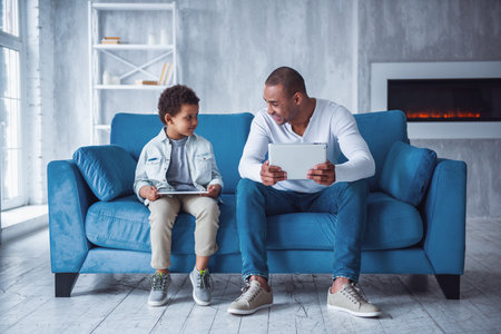 Happy father and son in casual clothes are using digital tablets and talking while sitting on couch at home, dad is smilingの写真素材