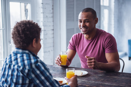 Handsome father and son in casual clothes are eating at the wooden table together at home, man is smilingの写真素材