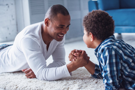 Happy father and son in casual clothes are arm wrestling and smiling while lying on the rug at homeの写真素材