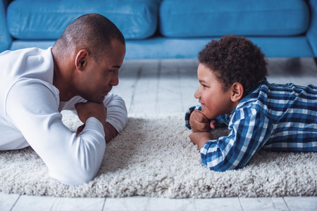 Happy father and son in casual clothes are looking at each other and smiling while lying on the rug at homeの写真素材