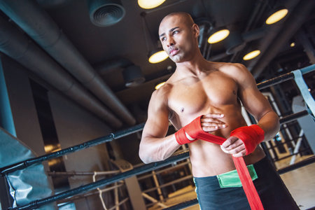 Handsome young boxer is wrapping hands with red bandage and looking away while preparing to train in gymの写真素材