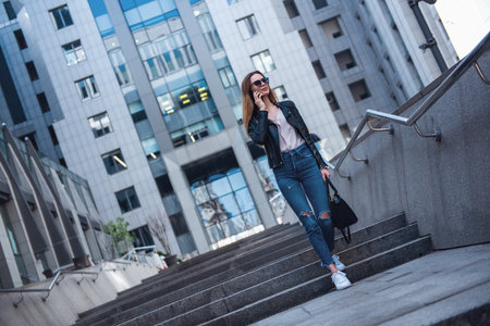 Full-length portrait of beautiful young girl in casual clothes and sun glasses talking on the mobile phone and smiling while walking in the city centerの写真素材