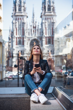 Beautiful young girl in casual clothes is holding a smartphone, looking upward and smiling while resting in city centerの写真素材