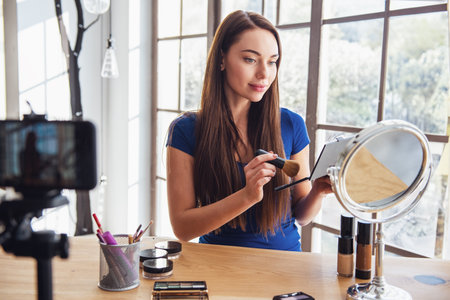Young beautiful woman blogger using cosmetics while shooting at camera at homeの写真素材