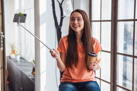 Young beautiful woman blogger while shooting selfie on phone using monopod sitting at window at home and drinking coffeeの写真素材