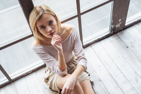 Portrait of young beautiful girl sitting on floor near big panoramic window and smile at cameraの写真素材