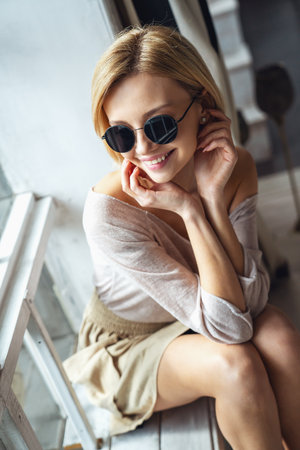 Portrait of young beautiful girl sitting on windowsill in sunglasses and smiling at camera at homeの写真素材