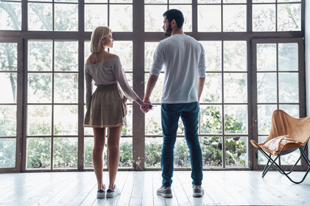 Young happy loving couple guy and girl standing in front of big panoramic window, back to camera holding handsの写真素材