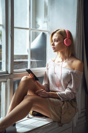 Young beautiful girl sitting on the windowsill and listening to music in pink headphonesの写真素材