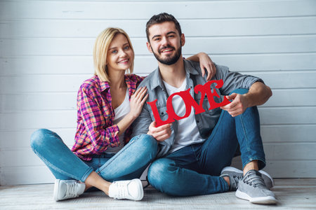 Young happy loving couple sitting on floor and holding sign "Love", smiling at cameraの写真素材