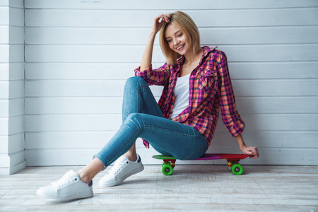 Portrait of young cheerful beautiful blonde girl sitting on skateboard and smiling at camera on light backgroundの写真素材