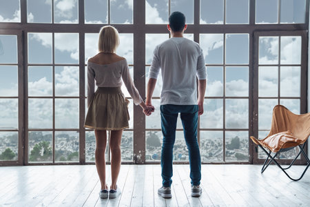 Young happy loving couple guy and girl standing in front of big panoramic window, back to camera holding handsの写真素材