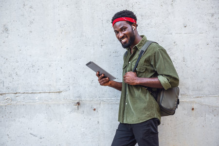 Young man listening to music on headphones using an electronic tablet while walking the streets of the bridge with a backpack near a large wallの写真素材