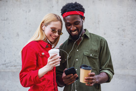 Young cheerful couple, where a man and a blonde woman stand on a background of a gray wall, listening to music on headphones and drinking coffeeの写真素材
