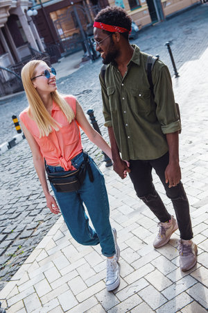Young cheerful couple where a man and a blonde woman are smiling holding each other's hands while walking down a city streetの写真素材