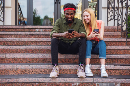 Young cheerful couple where man and blonde woman use their phones sitting on the stairs while walking around the cityの写真素材