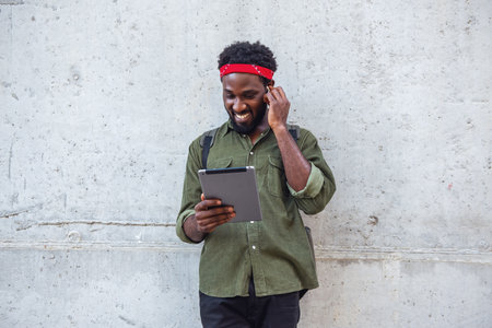 Young man listening to music on headphones using an electronic tablet while walking the streets of the bridge with a backpack near a large wallの写真素材
