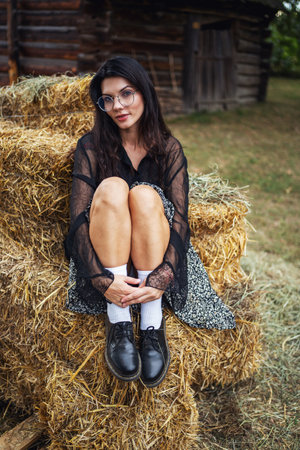 Retro style. A young beautiful girl with black chic hair in a black dress is posing near a haystack on the background of an old wooden house.の写真素材