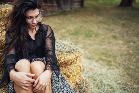 Retro style. A young beautiful girl with black chic hair in a black dress is posing near a haystack on the background of an old wooden house.の写真素材