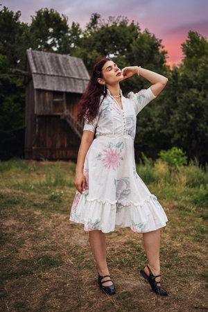 Retro style. Young beautiful girl with black chic hair in a white dress on the background of a forest, a mill and a red sunset.の写真素材