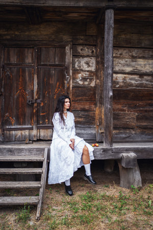 Retro style. Young beautiful sexy girl with black chic hair in white dress sitting on wooden stairs near wooden old house.の写真素材