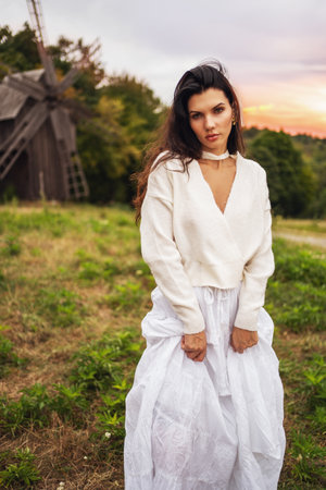 Retro style. Portrait of a young beautiful girl with black chic hair in a white dress on the background of a forest, a mill and a red sunset.の写真素材
