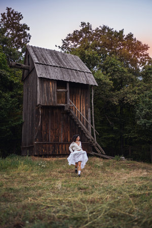 Retro style. Young beautiful girl with black chic hair in a white dress on the background of a forest, a mill and a red sunset. Runs towards the camera.の写真素材