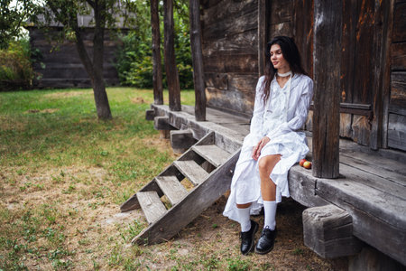 Retro style. Young beautiful sexy girl with black chic hair in white dress sitting on wooden stairs near wooden old house.の写真素材