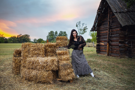 Retro style. A young beautiful girl with black chic hair in a black dress is posing near a haystack on the background of an old wooden house.の写真素材