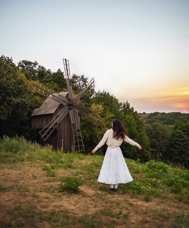 Retro style. Young beautiful girl with black chic hair in a white dress on the background of a forest, a mill and a red sunset. Stands with his back to the camera.の写真素材