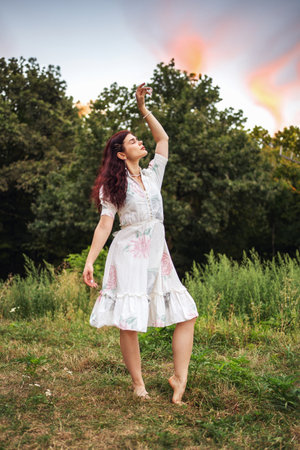 Retro style. Young beautiful girl with black chic hair in a white dress on the background of a forest and a red sunset.の写真素材