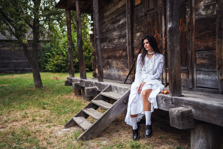 Retro style. Young beautiful sexy girl with black chic hair in white dress sitting on wooden stairs near wooden old house.の写真素材