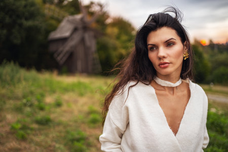 Retro style. Portrait of a young beautiful girl with black chic hair in a white dress on the background of a forest, a mill and a red sunset.の写真素材