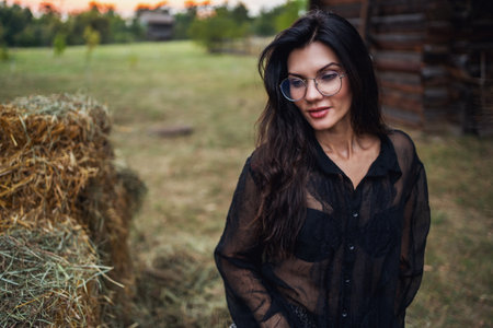 Retro style. A young beautiful girl with black chic hair in a black dress is posing near a haystack on the background of an old wooden house.の写真素材