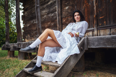 Retro style. Young beautiful sexy girl with black chic hair in white dress sitting on wooden stairs near wooden old house.の写真素材