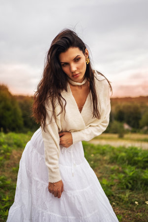 Retro style. Full-length portrait of a young beautiful girl with black chic hair in a white dress against a background of a forest and a red sunset.の写真素材