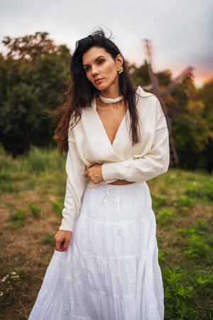 Retro style. Full-length portrait of a young beautiful girl with black chic hair in a white dress against a background of a forest and a red sunset.の写真素材