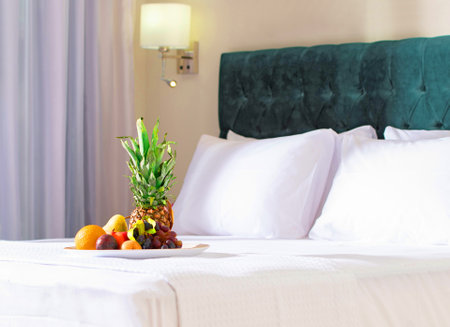Fruity welcome gift in hotel interior blur background. Close up of healthy breakfast fruit plate with pineapple on white bed linen in modern bedroom.の写真素材