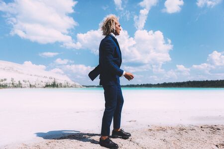 Blond curly man walking on the beach of crystal clear sea. Dressed business suit, blue color. Summer, trendy. Travel topicの写真素材