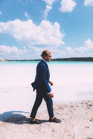 Blond curly man walking on the beach of crystal clear sea. Dressed business suit, blue color. Summer, trendy. Travel topicの写真素材