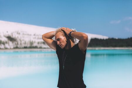 Curly blond man posing at the clear beach. Dressed all black. Awesome background, clear water and sky. Trendy, tropical and attractive man. Summer time.の写真素材