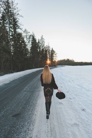 Young and alone girl dressed fur coat and dress standing around outdoor road. Sunset on the background. Sunlight and faded colors. Snowの写真素材