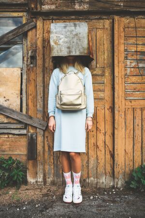 A long haired blonde woman, wearing a powder blue dress, white socks with pink marijuana motifs, white trainers and a pink backpack the wrong way round, wearing on her head a rusty metal containerの写真素材