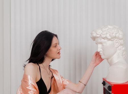 Attractive skinny girl in a loose robe and lingerie wants to warm up near the heater and looks at the head of a statue.の写真素材
