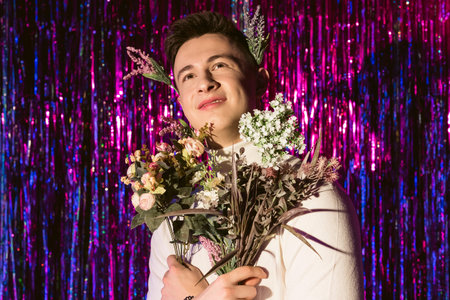 Portrait of a young man in white turtleneck sweater holding bouquet of flowers standing against shiny ribbons background.の写真素材
