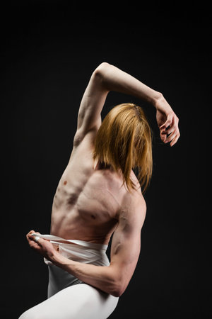 Young athletic professional ballet dancer with a bare torso and white dance tights is in perfect shape and posing with bright yellow slippers in his hands over a black background.の写真素材