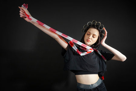 Portrait of a poor, skinny girl in black, rumpled clothes and a strange hat that made of toilet paper tubes wrapper in a traffic or caution tape, isolated on a black background.の写真素材