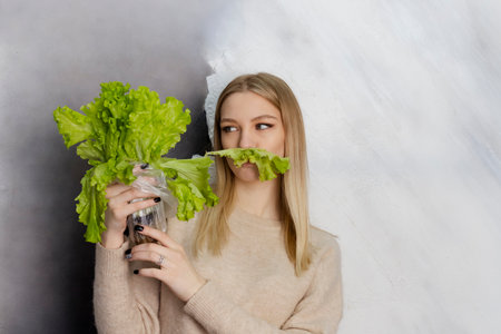 A beautiful young blonde in light clothing holds lettuce leaves in her hands against a light background. lettuce leaves mustache.の写真素材