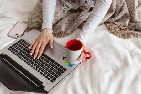 A beautiful girl sitting on a bed wrapped in a blanket with a warm drink in a mug and working on a laptop.の写真素材
